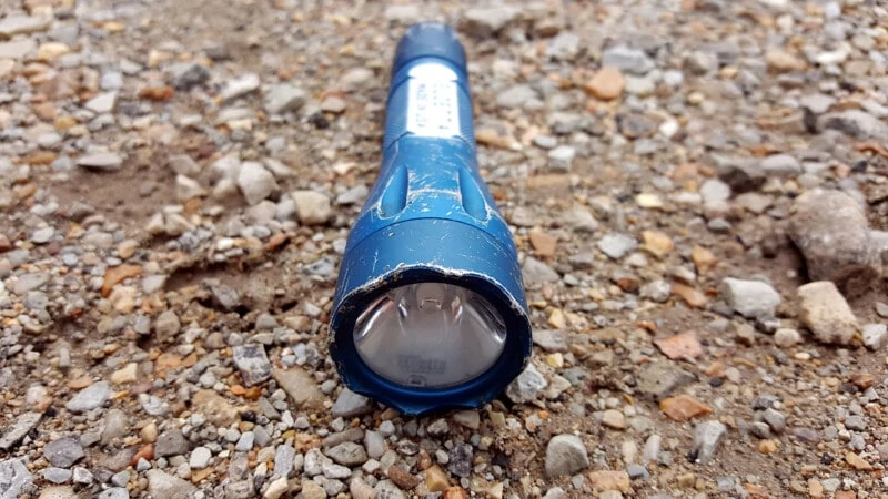Blue Elzetta Bones lying on gravel, showing wear on type II blue anodizing. Several scratches and nicks in the surfaces of the flashlight.