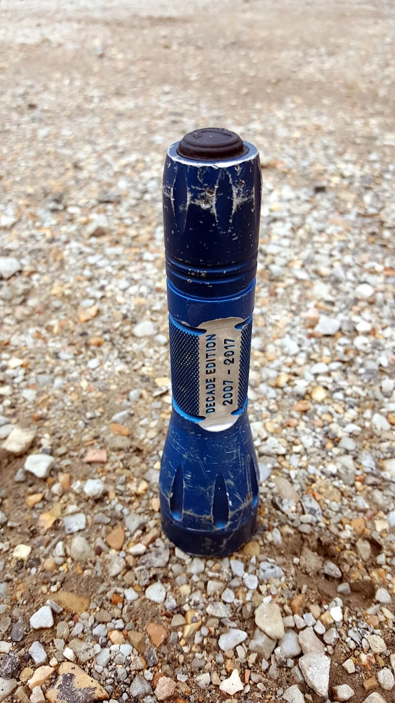 Blue Elzetta Bones standing vertical on gravel, showing wear on type II blue anodizing. Several scratches and nicks in the surfaces of the flashlight. Text on light: "Decade Edition 2007 - 2017"