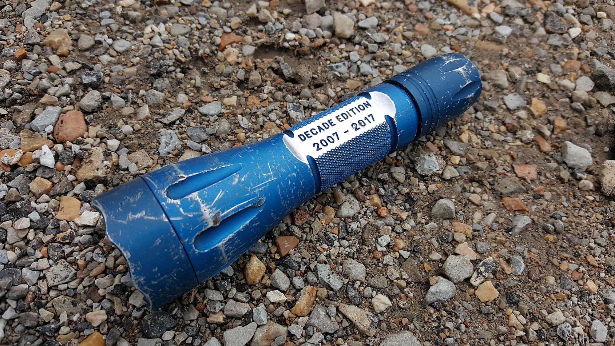 Blue Elzetta Bones lying on gravel, showing wear on type II blue anodizing. Several scratches and nicks in the surfaces of the flashlight. Text on light: 